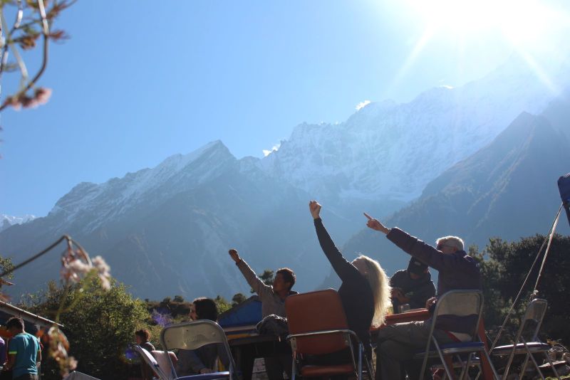 The picture is of 5-6 people sitting on a chair with table. They are sitting Infront of the mountainous landscape and are pointing to something  which is not in the photoo. 